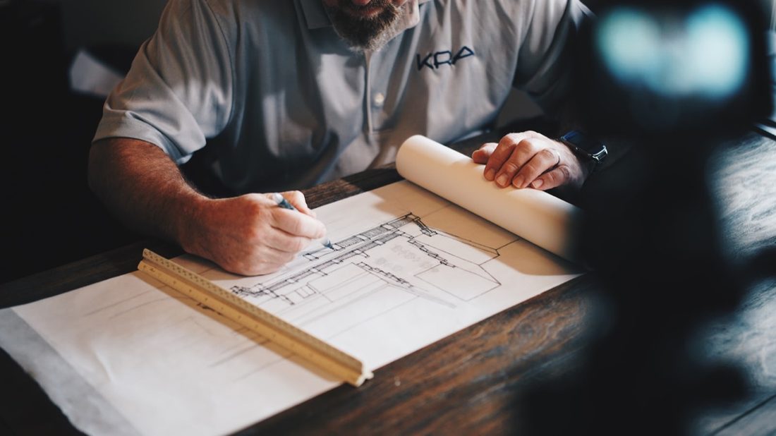Hands drawing a building plan in pencil with a scale ruler on the desk